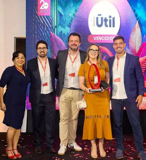 Pessoas em um evento de premiação, com um fundo exibindo a marca "Banco Útil" e a palavra "Vencedor". A mulher ao centro segura um troféu, representando o prêmio recebido no evento Cantarino Brasileiro.