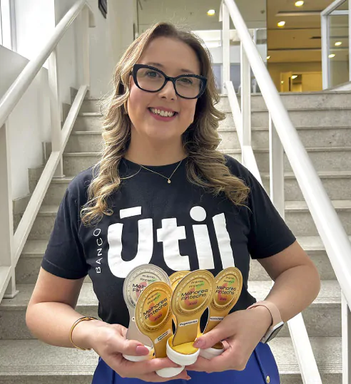 Mulher sorridente segurando prêmios da Finatech, vestindo uma camiseta preta com a marca "Banco Útil", em uma escadaria de um ambiente corporativo.